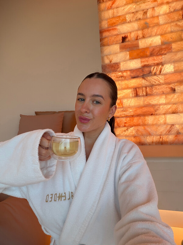 Emily in a white spa robe holding a cup of tea by a salt wall